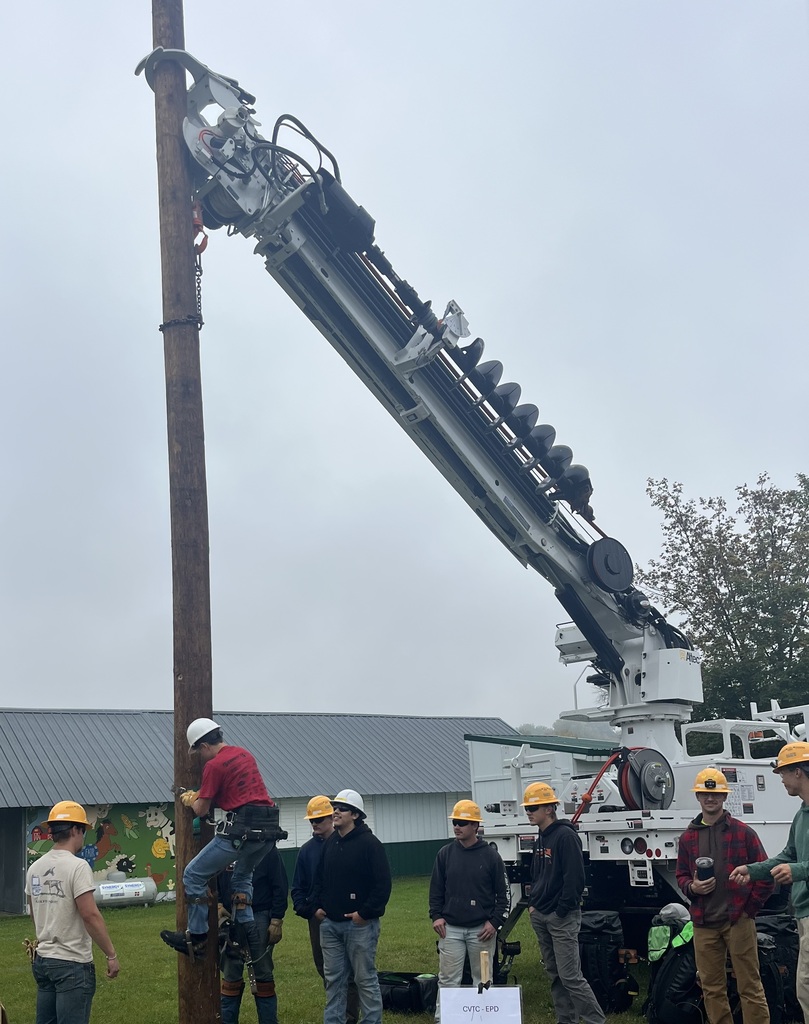 A high school student practicing climbing a wooden utility pole wearing climbing gear, while a group of students in hard hats and safety gear watch alongside a large utility crane truck outdoors.