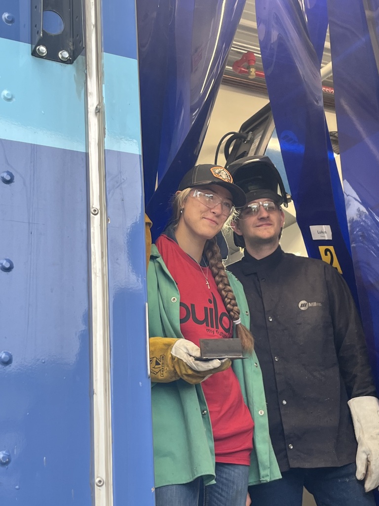 Two high school students wearing safety glasses and protective gear standing inside a mobile welding display trailer. One student wears a red "Build My Future" t-shirt, green welding jacket, and work gloves while holding up a welded piece of metal, next to another student in a black welding coat.