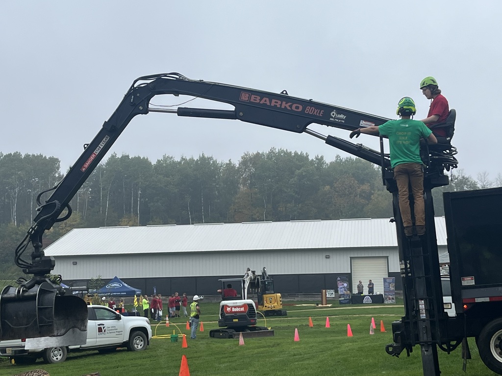 A high school student operating a large Barko heavy machinery claw arm outdoors at an event field, while a supervisor stands on the platform giving guidance.