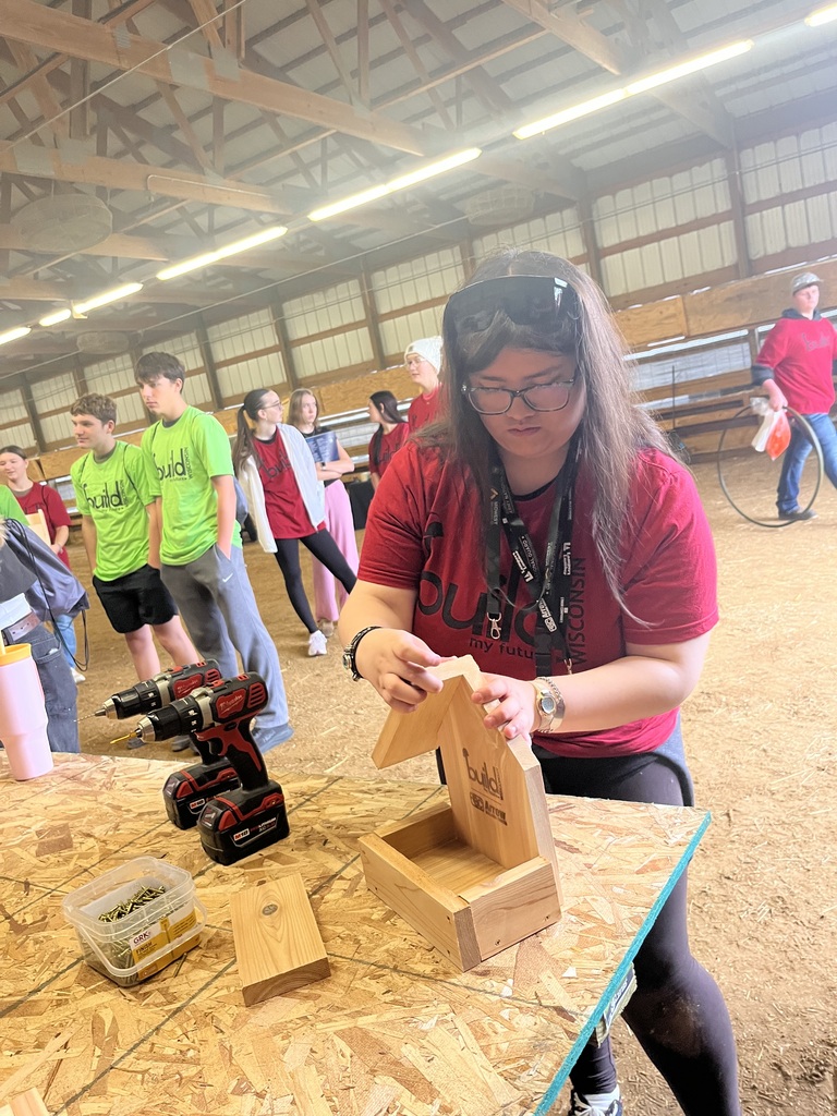 female high school student wearing a red "Build My Future" shirt, safety glasses, and lanyard assembling a small wooden birdhouse using screws at an indoor workbench, with power drills and a container of screws nearby.