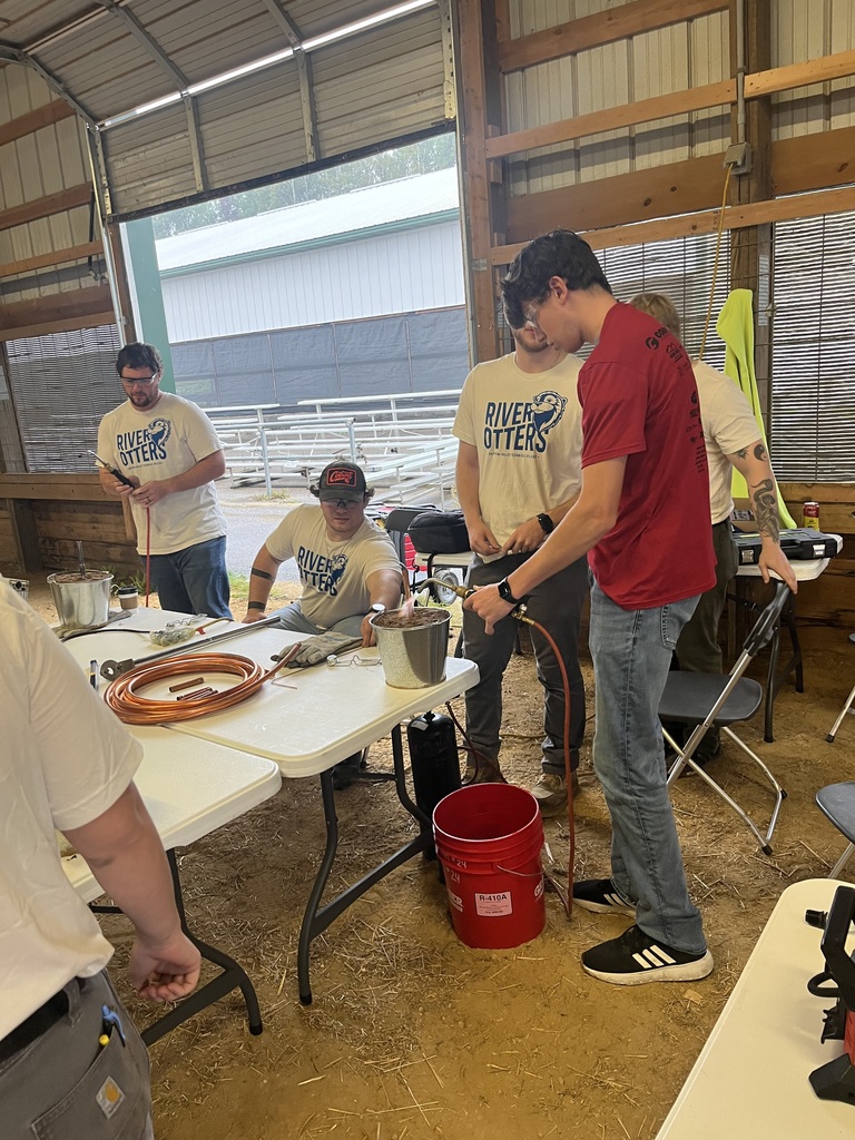igh school students wearing safety glasses practicing copper pipe soldering using a blowtorch at an indoor table, supervised by instructors in matching t-shirts.