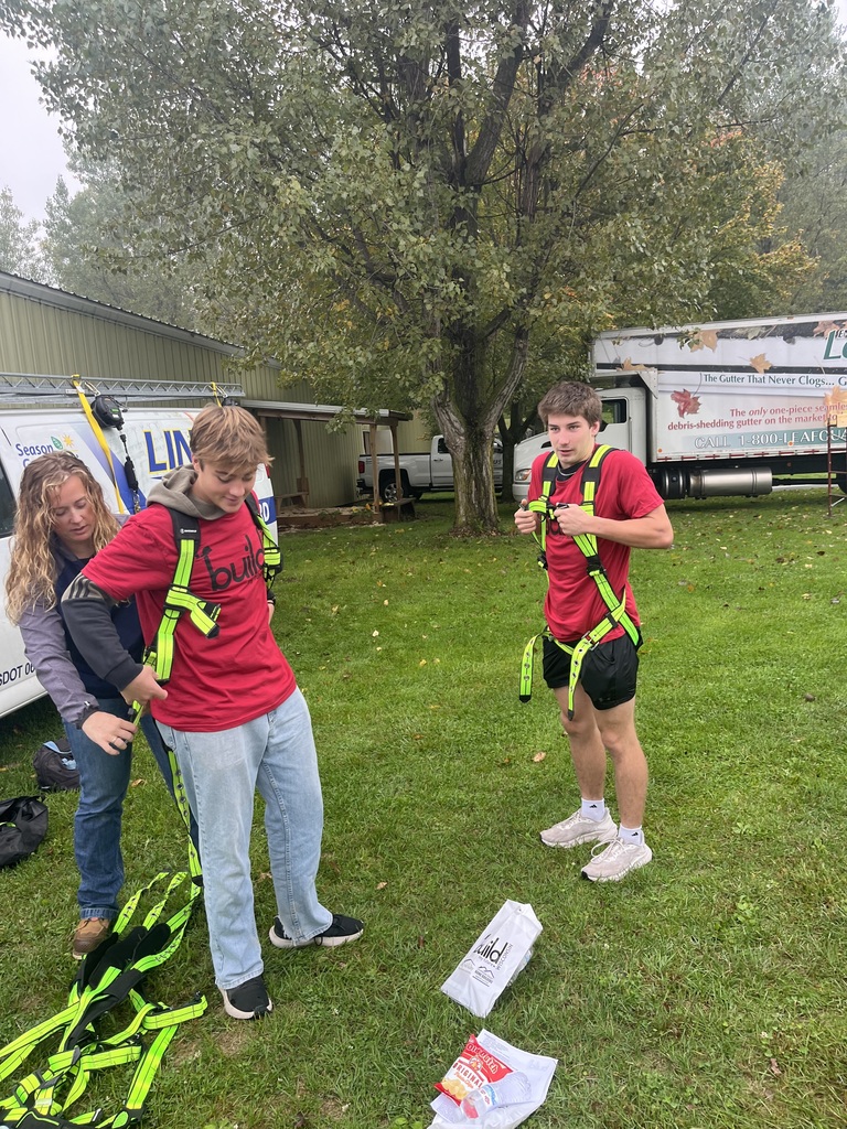 wo male high school students in red "Build My Future" t-shirts trying on bright neon yellow safety harnesses with the assistance of an instructor outdoors near work vehicles.