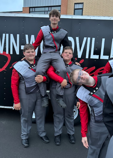 Five high school marching band percussion members in grey and red uniforms pose playfully outdoors. One student sits high on the shoulders of two bandmates, while another leans into the frame with a goofy expression and eyeglasses