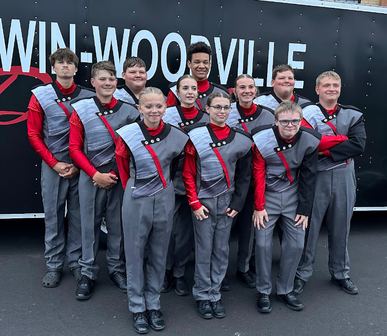 Eleven high school marching band percussion members in grey and red uniforms pose together outdoors in front of a trailer labeled "BALDWIN-WOODVILLE". They are lined up in two rows, smiling for the camera
