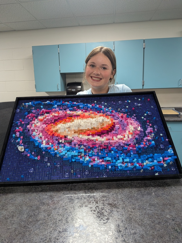 A smiling female student holding up a large, framed colorful LEGO build depicting a galaxy.