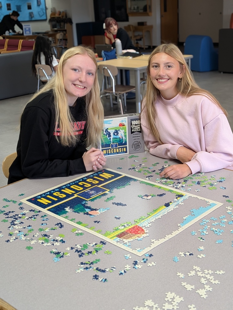 Two smiling female high school students sitting at a table together in a study hall, working on a large 1,000-piece "Wisconsin" jigsaw puzzle.