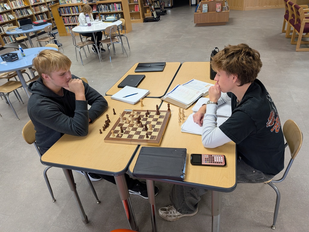 Two male high school students sitting at a desk across from each other, focused intently on a game of chess with notebooks and a calculator nearby.