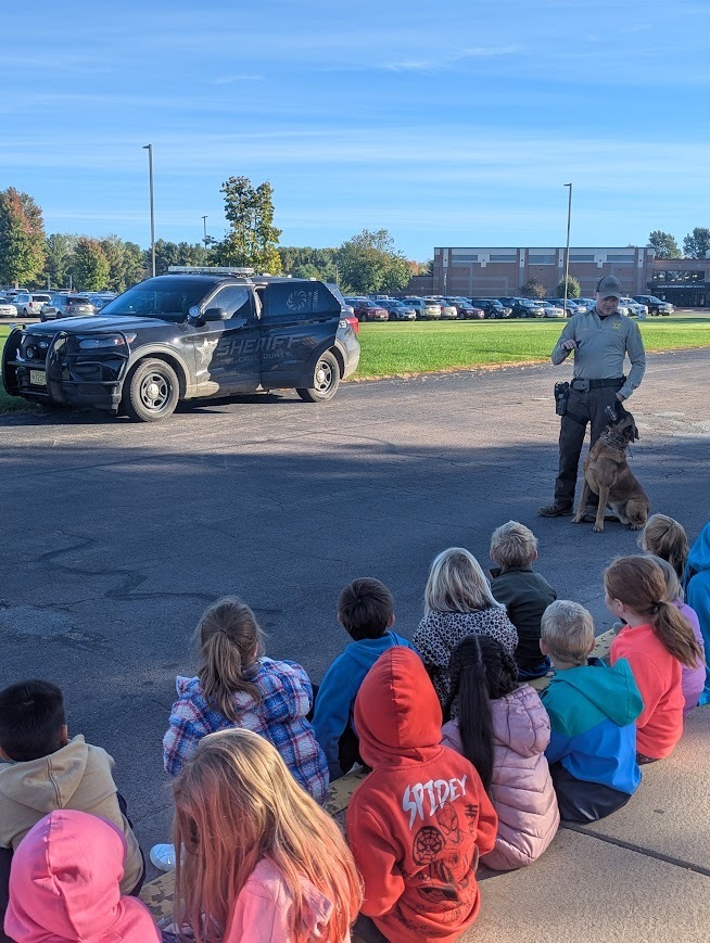 Kindergarten students sit attentively on the pavement as Deputy Jordan Smith stands beside a St. Croix County Sheriff’s vehicle and introduces them to K9 Aires