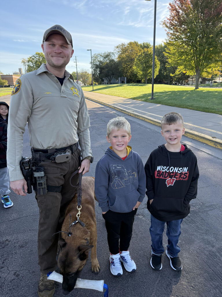 A close-up of two smiling kindergarten students (one in a Wisconsin Badgers hoodie) posing for a photo with Deputy Jordan Smith and K9 Aires, who is holding a training toy