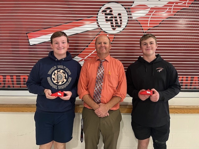 Three people standing side-by-side in front of a red and white Baldwin-Woodville wall graphic. On the left, Eli Lium holds a small red shoe award; in the center, Mr. Larson stands smiling in an orange dress shirt and tie; on the right, Adam Meier stands holding his matching red shoe award.