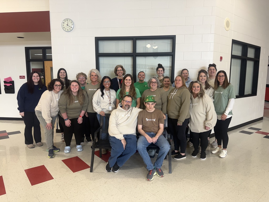 A group of school staff members smiling and standing together in a school hallway in front of a brick wall and window. In the center, a teenage boy wearing a green hat and a dark brown shirt sits next to a man. Several staff members around them wear matching light green and beige "Phelan Lucky" shirts to show their support.