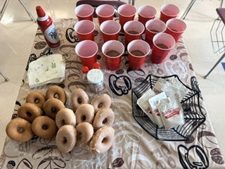 Table setup for a classroom celebration featuring glazed donuts, Swiss Miss hot cocoa packets in a wire basket, red cups, whipped cream, and sprinkles on a coffee-themed tablecloth.