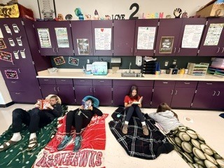 Four students lying on cozy blankets along the classroom wall, reading books during a morning reading celebration. One blanket prominently features the red and white "Blackhawks" text.
