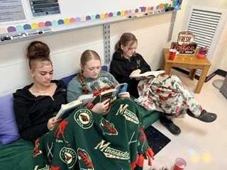 hree students sitting side by side wrapped in fleece blankets, intently reading their books in a classroom setup. A small wooden table nearby holds cups and a "Fresh Coffee House" sign.