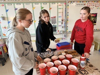 Three middle school students standing at a refreshment table lined with cups of hot cocoa, whipped cream, and pastries during classroom Fika time.