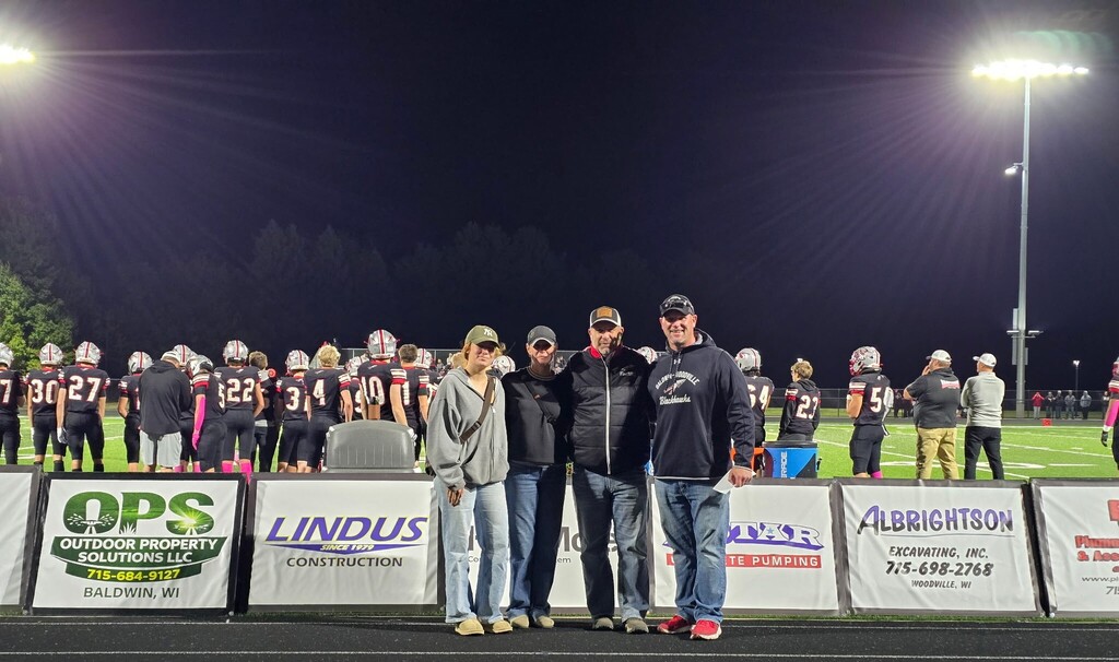 Maci Swanson, Heather Swanson, Jeff Swanson, and Jason Sell smiling together on the athletic track under stadium lights during a football game.