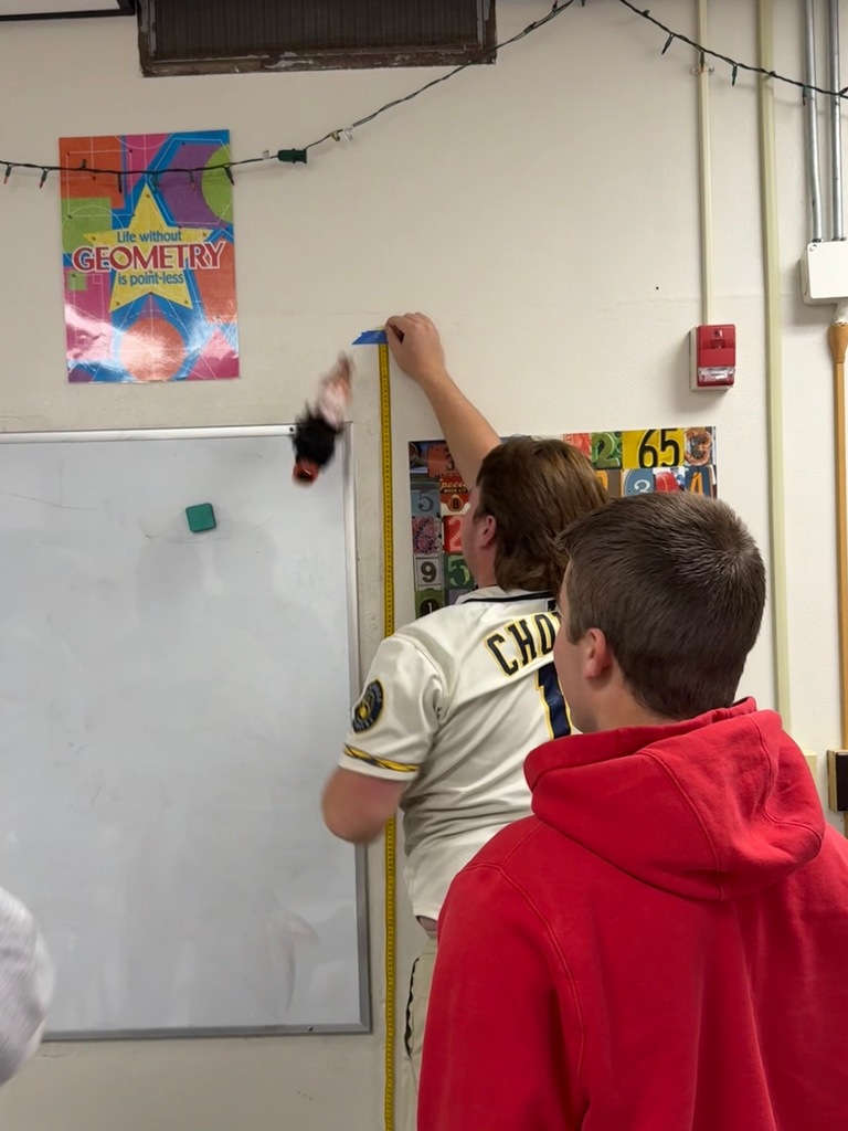 Students watching intently as a doll attached to rubber bands bounces near a measuring tape on a classroom wall during a statistics experiment.