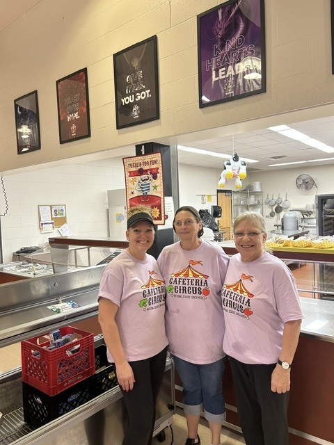 Three kitchen staff members wearing matching light purple "Cafeteria Circus" t-shirts smiling together behind the cafeteria serving line.