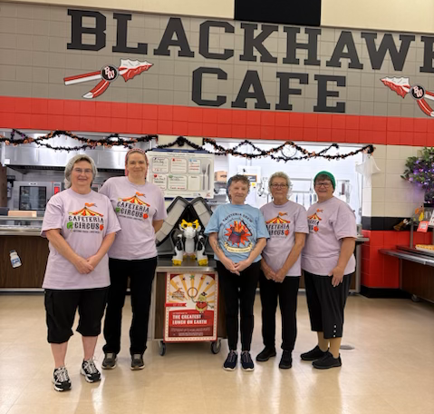 Five cafeteria staff members wearing matching lavender "Cafeteria Circus" t-shirts posing together under the Blackhawk Cafe sign in the school lunchroom.