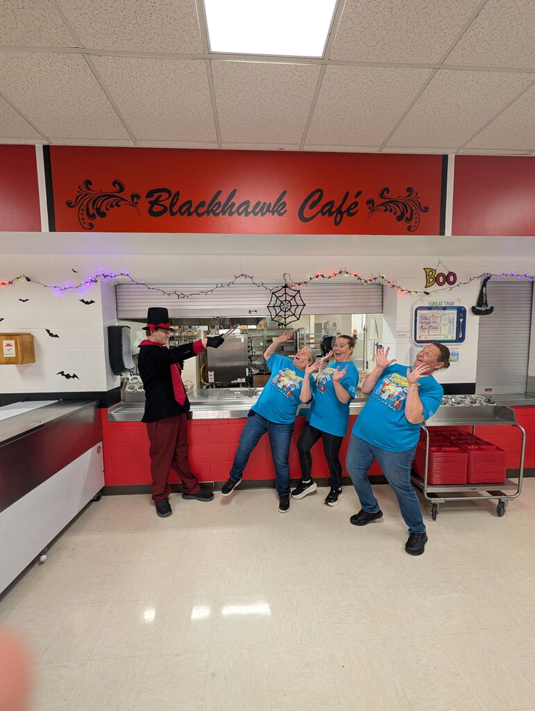 High school cafeteria staff members wearing matching blue shirts striking fun, dramatic poses in front of the Blackhawk Café serving counter alongside a staff member dressed as a magician.