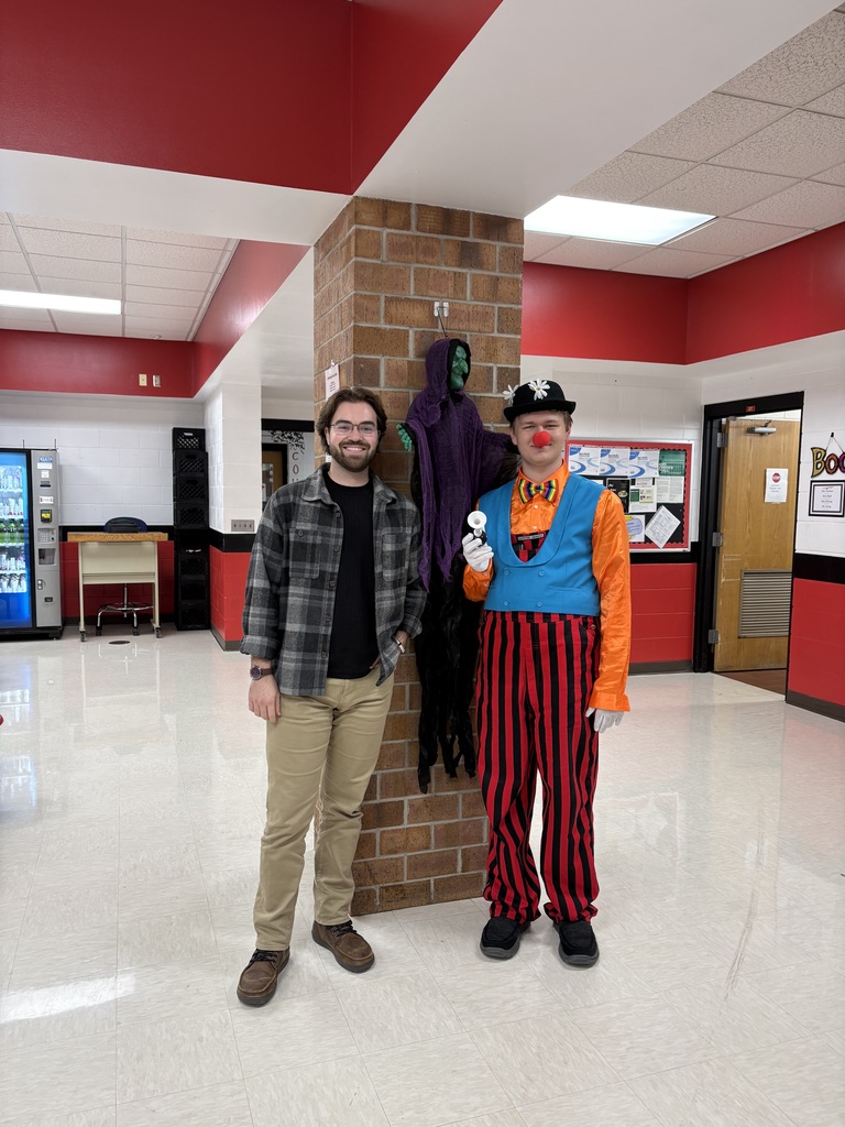 A staff member and a student dressed in a colorful circus clown costume with striped pants and a red nose posing together in the high school hallway