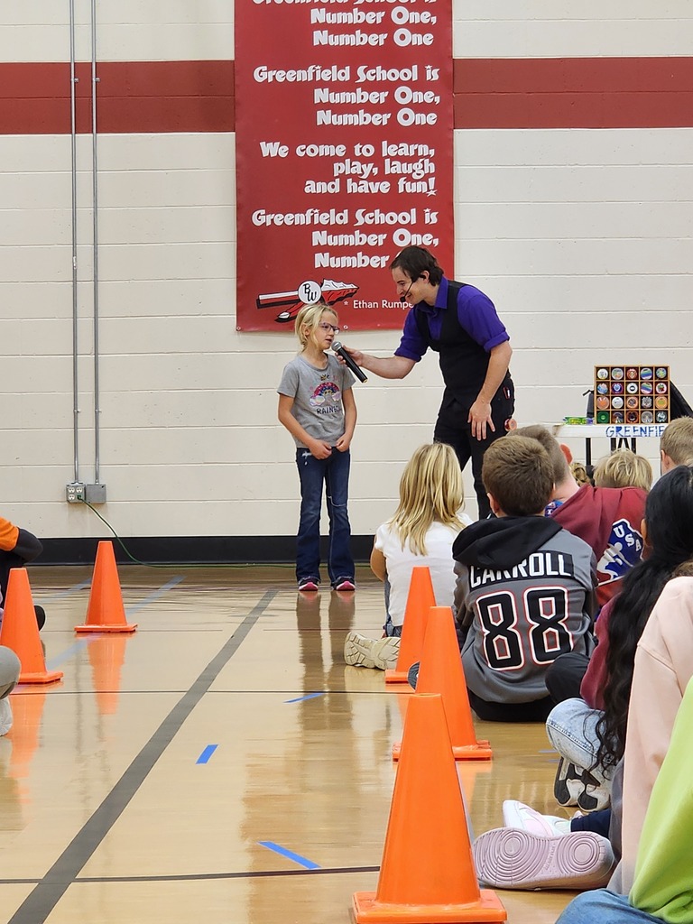 The assembly performer holding a microphone to a student volunteer on stage in the gymnasium in front of a banner reading "Greenfield School is Number One".