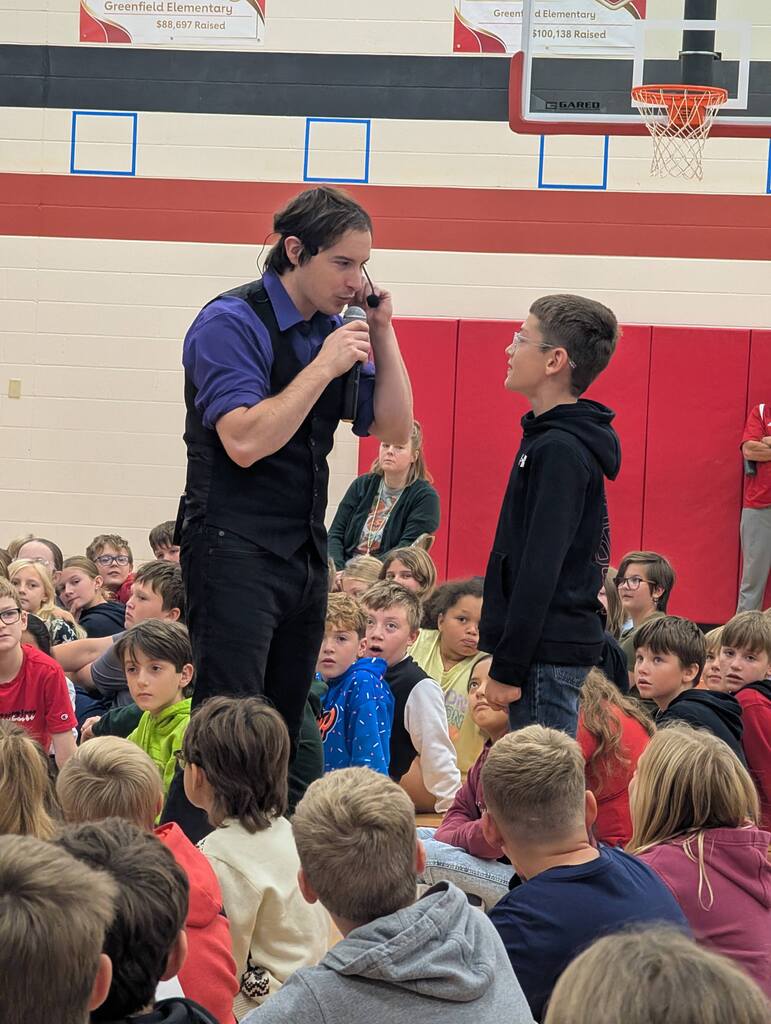 The NED Show performer wearing a purple shirt and black vest interacts directly with a student holding a microphone in front of a seated elementary assembly audience.