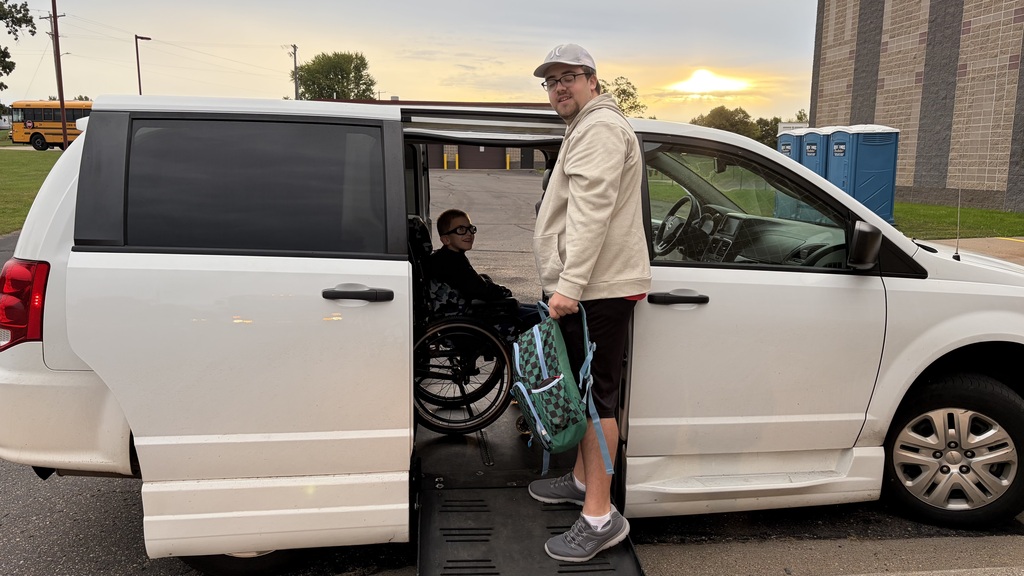 A Baldwin-Woodville school district employee holding a backpack and smiling next to an accessible van ramp while a student in a wheelchair looks out from inside the van during a morning drop-off.