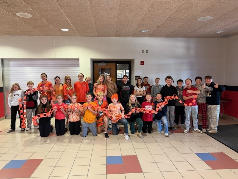 A large group of middle school students posing in a hallway, holding up a long, continuous chain made of orange paper links