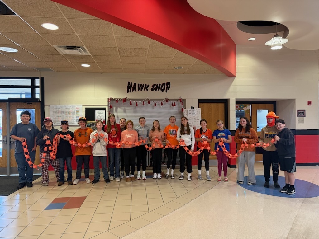 A line of middle school students standing together in a school hallway beneath a sign reading "Hawk Shop," collaboratively holding up a long connected chain of orange paper links stretched across the room.