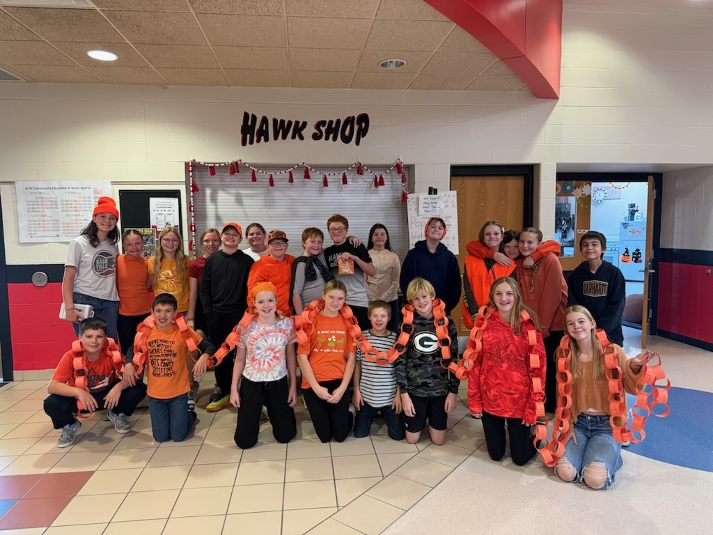 A group of middle school students wearing orange clothing and posing together in front of the "Hawk Shop" storefront in a school hallway, with several students draped in long orange paper chains