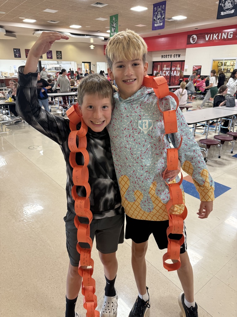 Two smiling young boys posing together in a school cafeteria with long orange paper chains draped around their shoulders