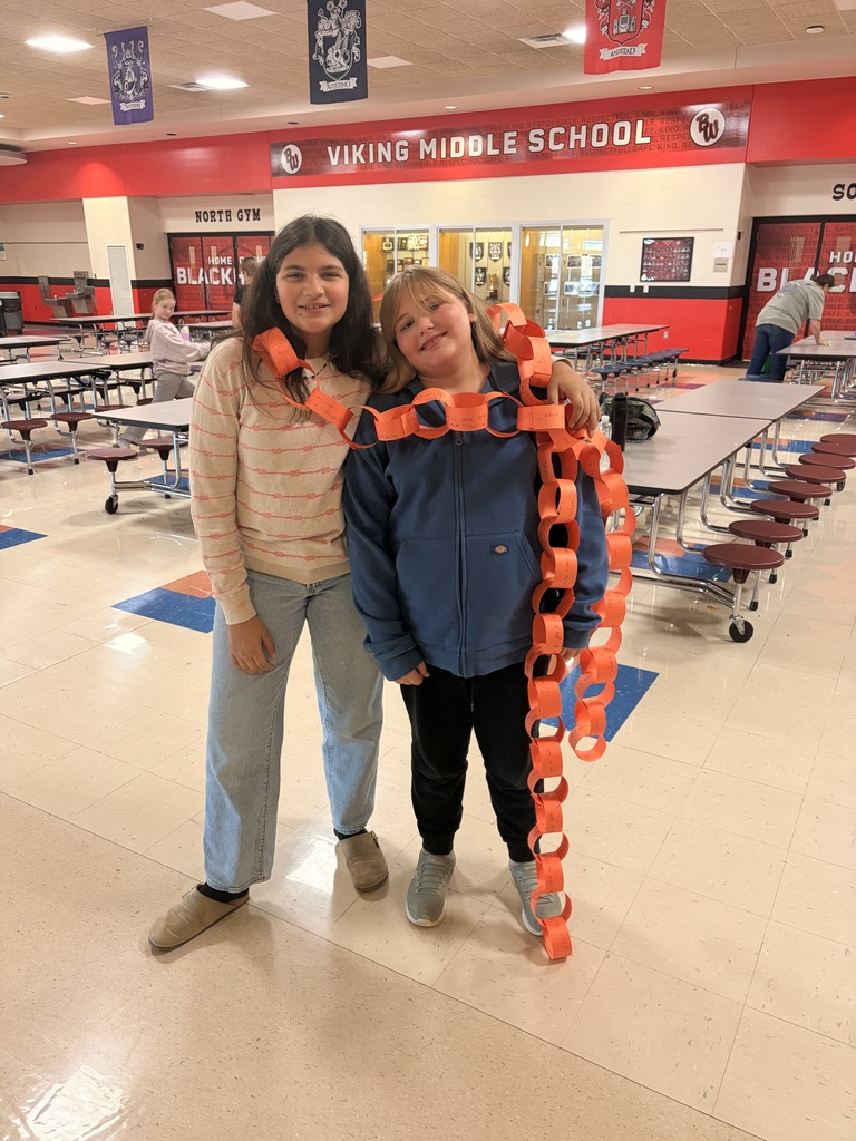 Two middle school girls smiling in a school cafeteria, wrapped together in a long orange paper chain