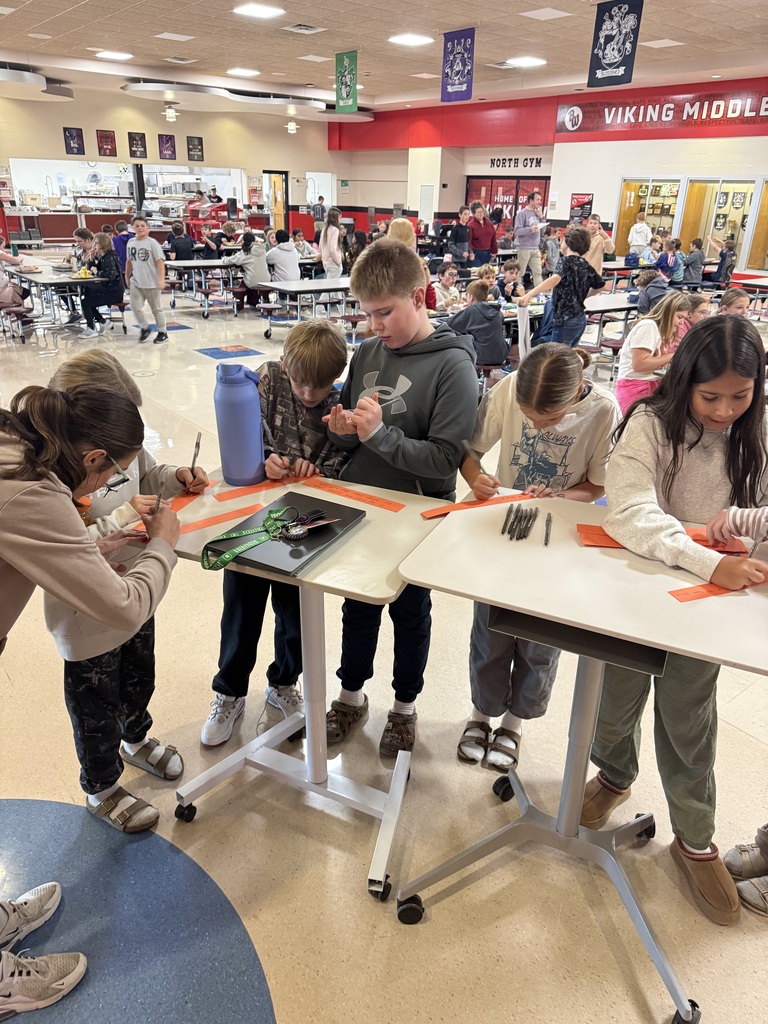 Five middle school students standing at white rolling tables in a school cafeteria, using markers to write messages on strips of orange paper.