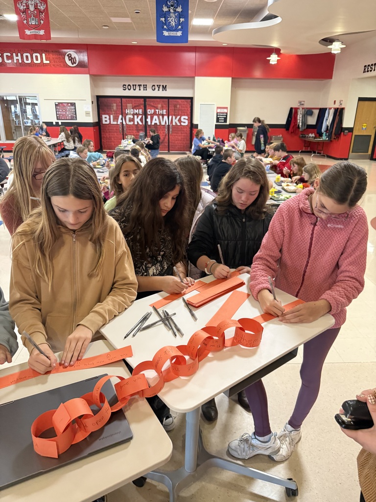 A group of middle school girls gathered around a table in a school cafeteria, writing messages on strips of orange paper and looping them together into an orange paper chain.