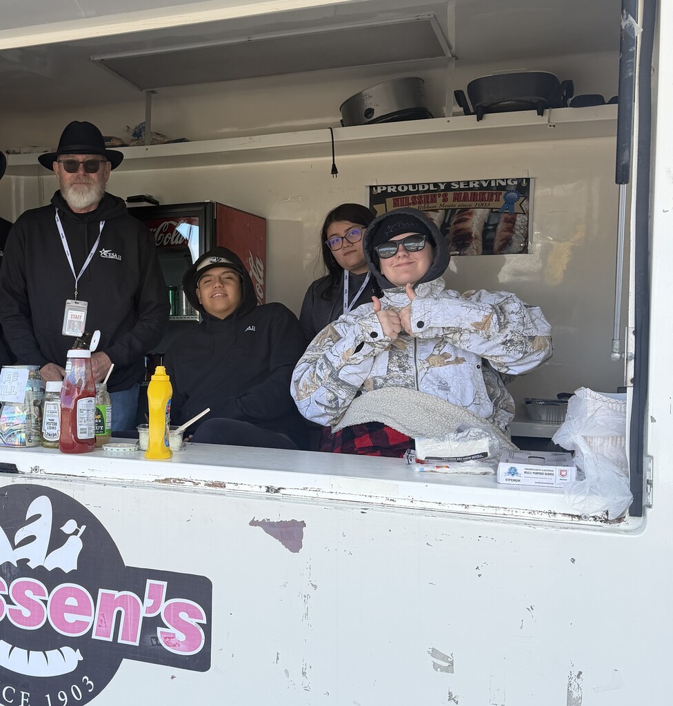 Four people smiling inside a white food truck serving stand, with condiments on the counter and a "Nilssen's Market" sign on the back wall.