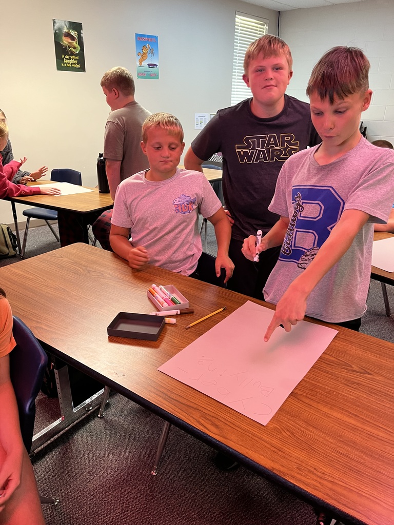 3 boys standing at a table pointing a blank sheet of paper
