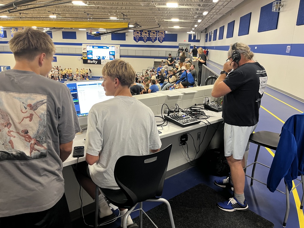 Two students work at a computer while an adult operates audio equipment and wears a headset at the media station overlooking a volleyball match in the school gym.