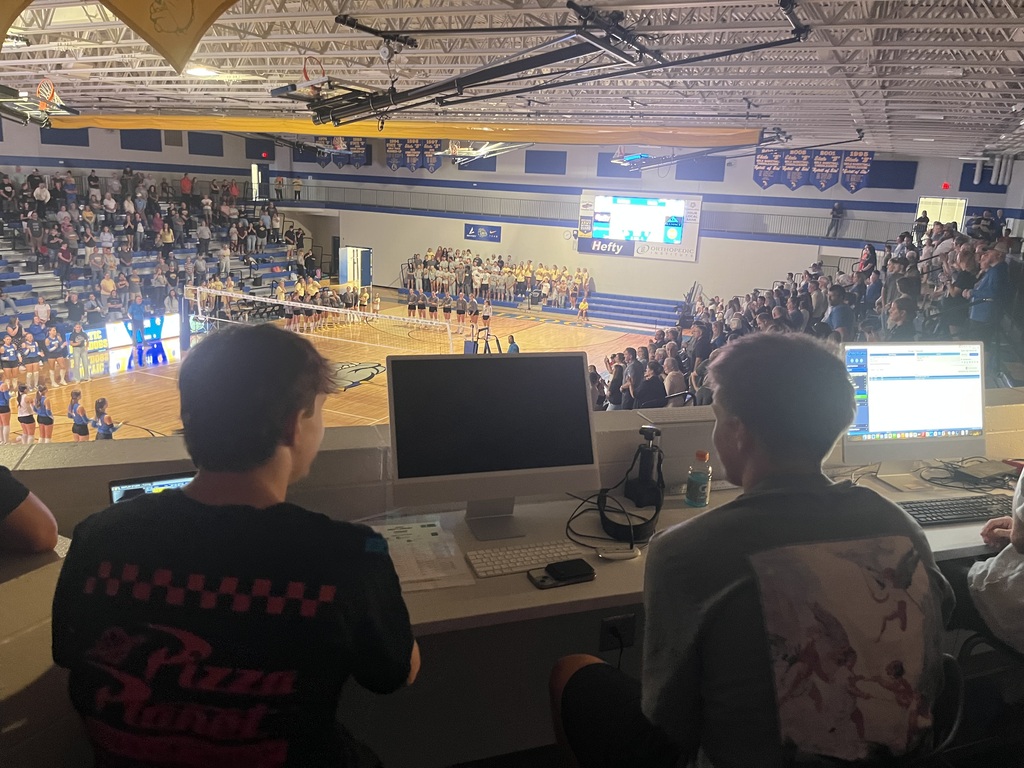 Two students operate computers from the media station above the gym while a volleyball match takes place on the court below in front of a large crowd.
