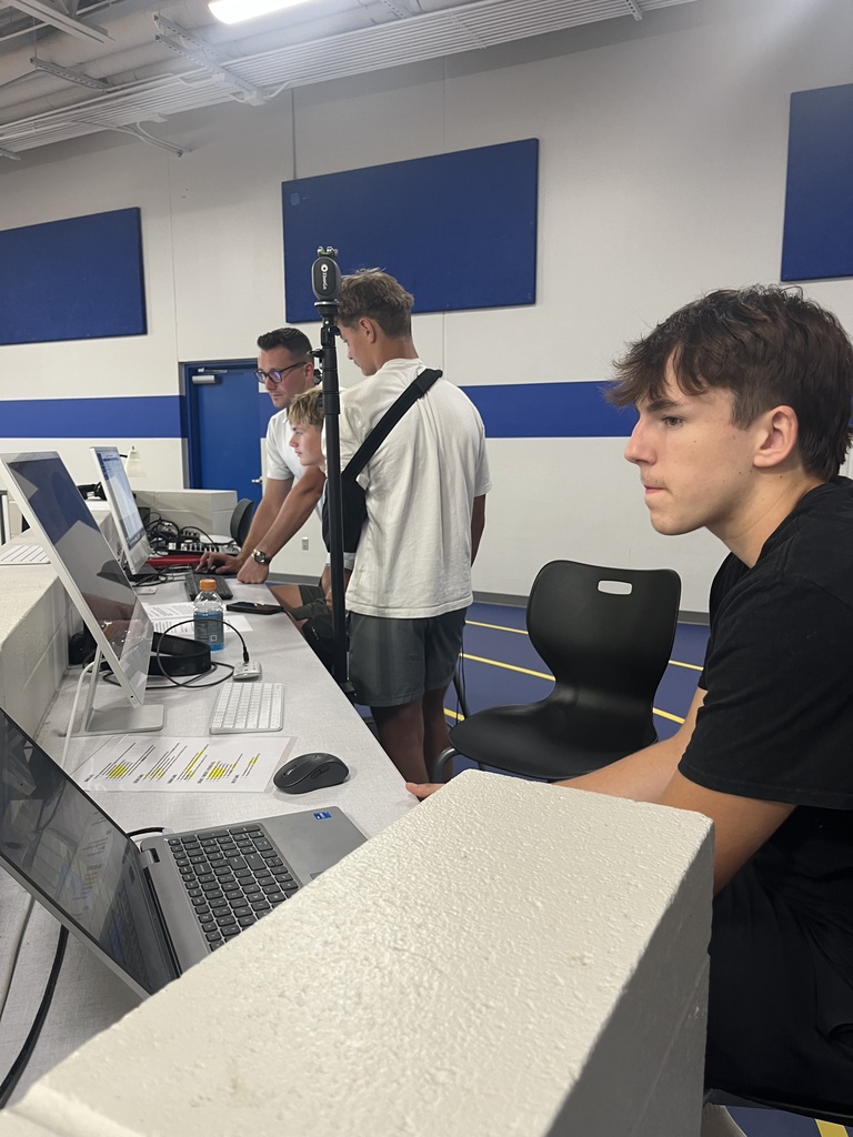 A student focuses on a laptop at the gym media station while other students and an adult work with computers and production equipment nearby.
