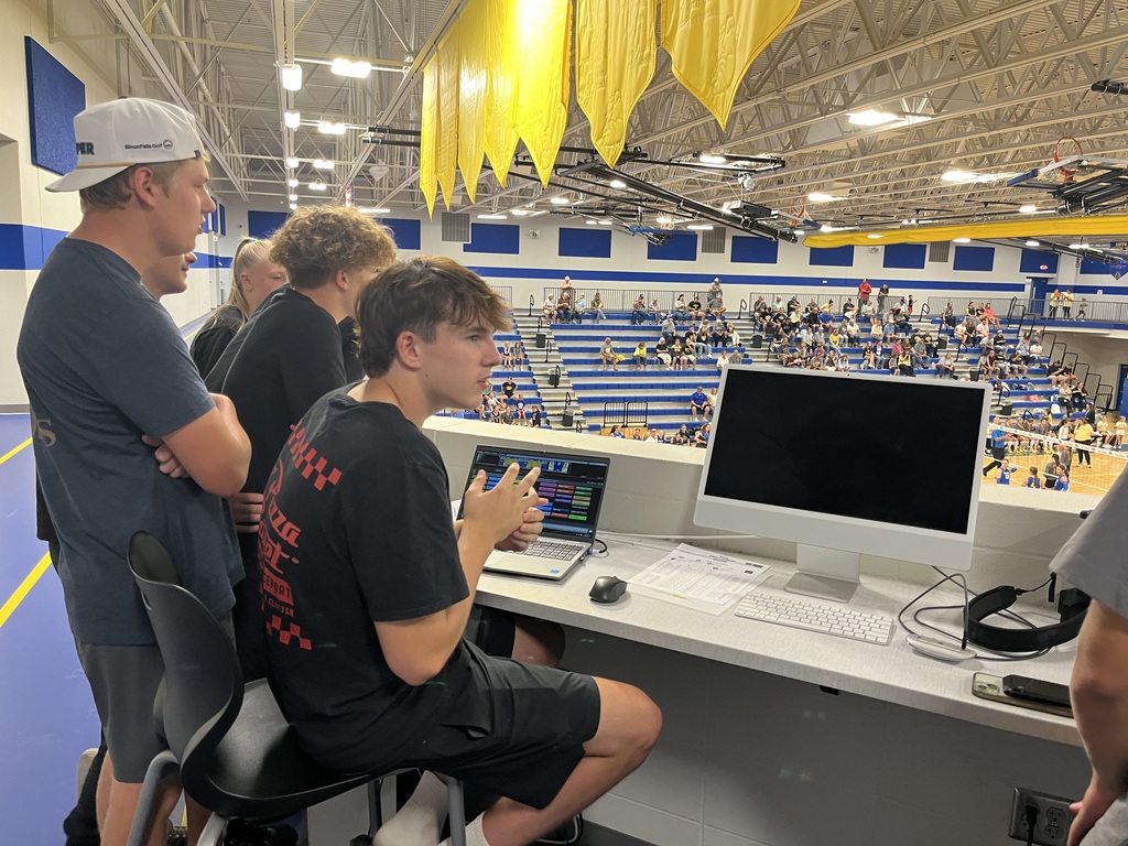 Students work together at the media station overlooking the school gym, using computers and production equipment during a volleyball match.