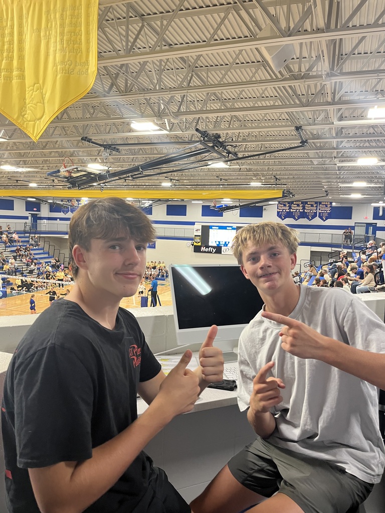 Two high school students work at the media station overlooking a volleyball match in the school gym. One gives a thumbs-up while the other points toward him.