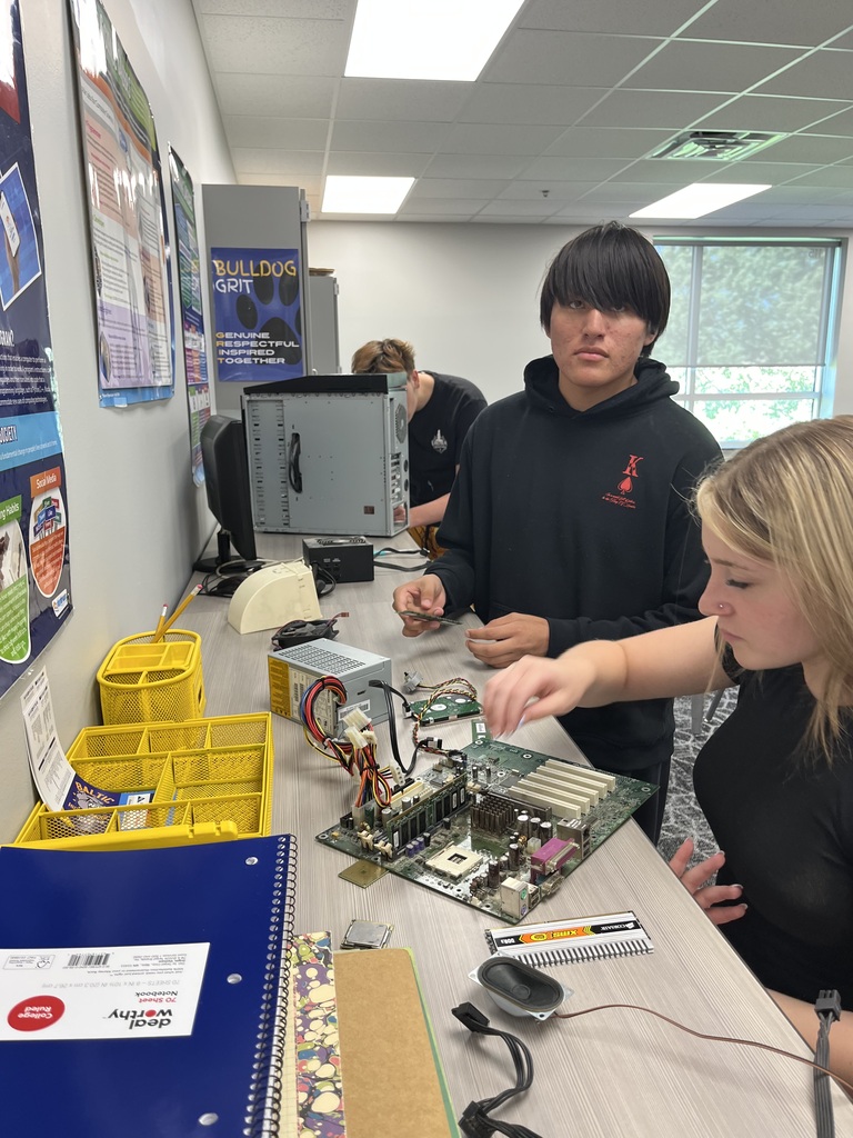 Three students collaborate around a disassembled desktop computer, examining a motherboard and other internal components during a hands-on computer science activity.