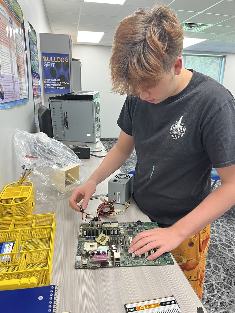A student works with a disassembled computer motherboard, connecting power supply cables and examining internal computer components during a hands-on computer science lesson.