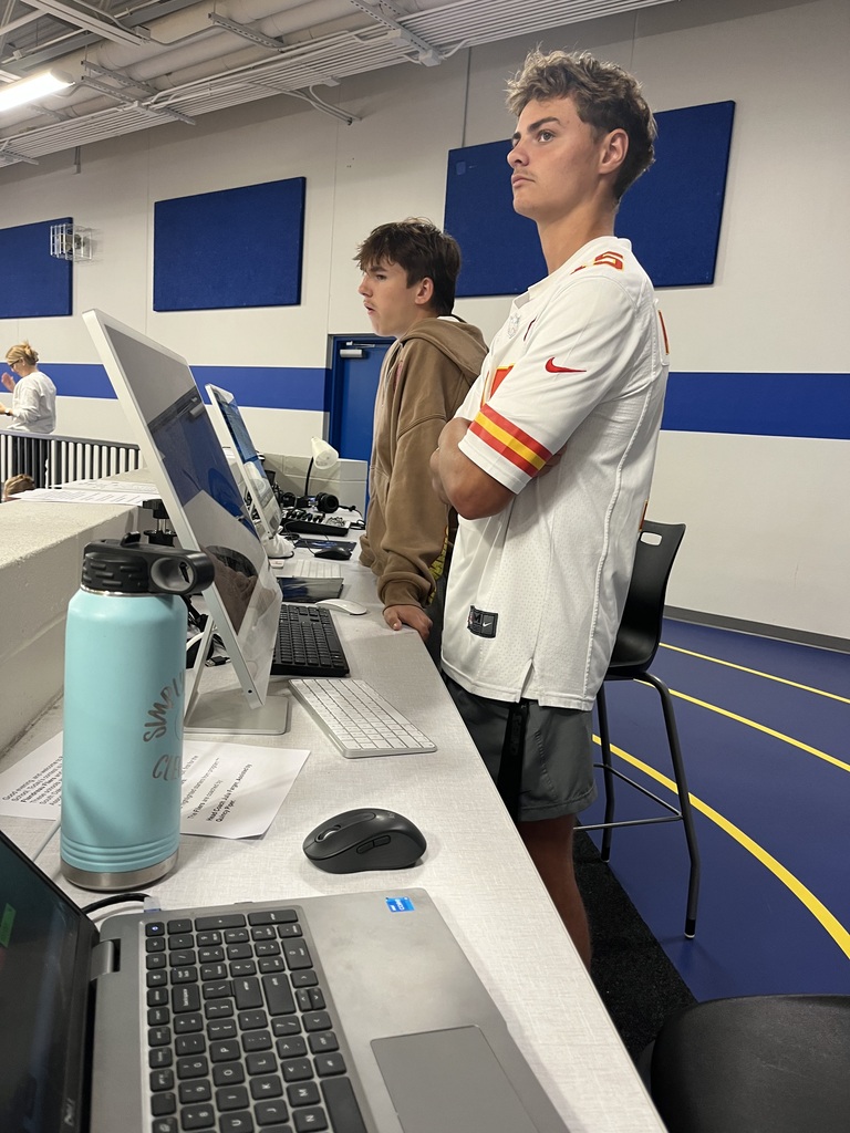 Two students stand at the media and production station overlooking the school gym, preparing to operate computers and equipment during a school activity.