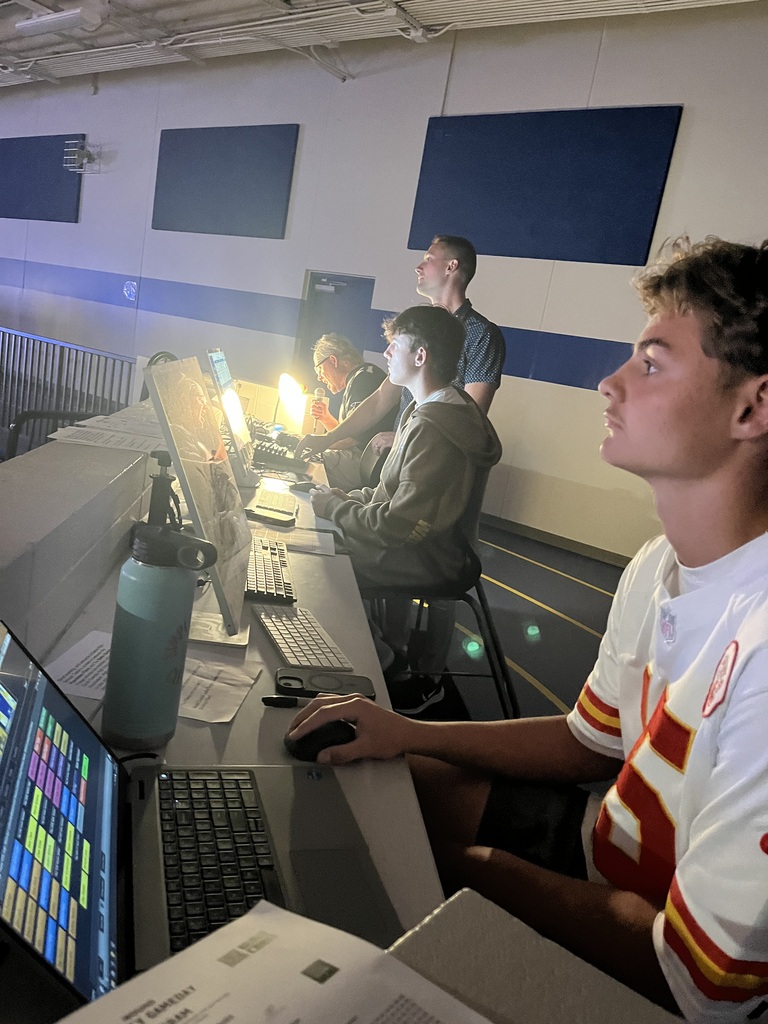 Students and an adult work together at the gym media station, using computers and production equipment to support a school event.