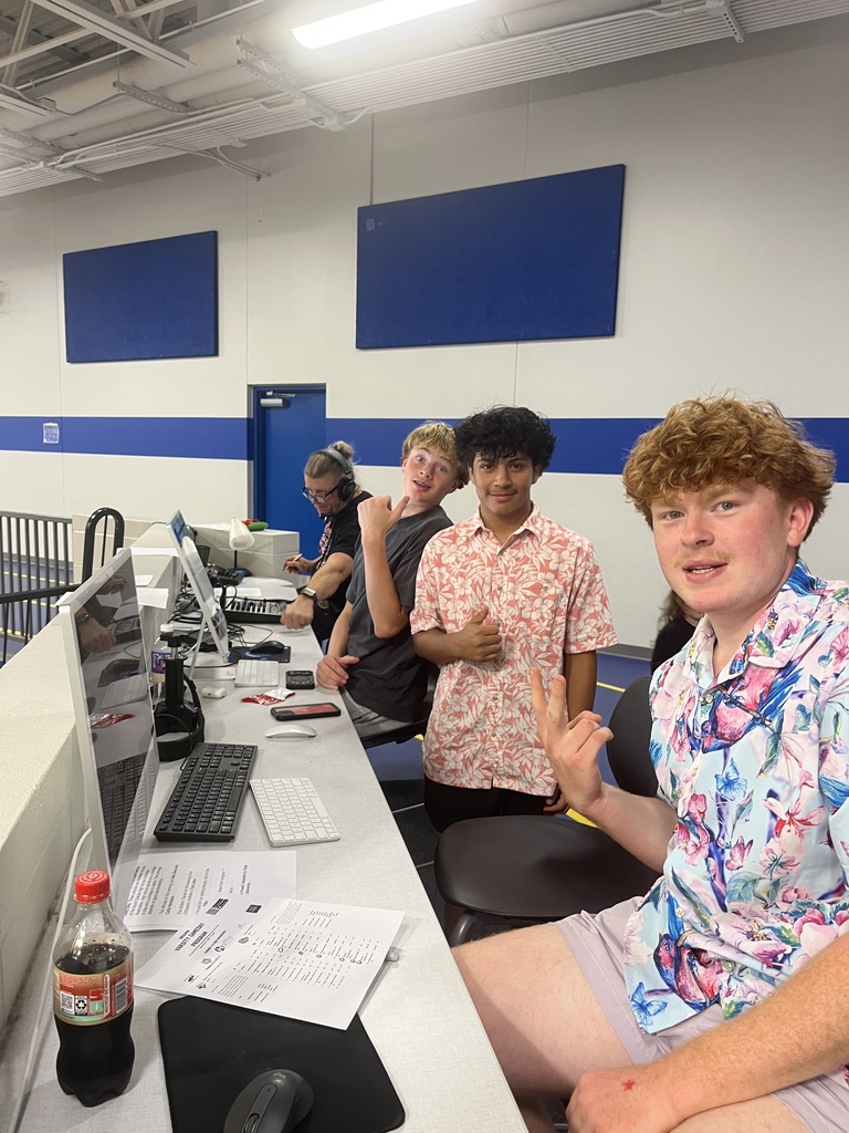 Three students pose together at the media station while an adult works with audio and computer equipment in the background during a school event.
