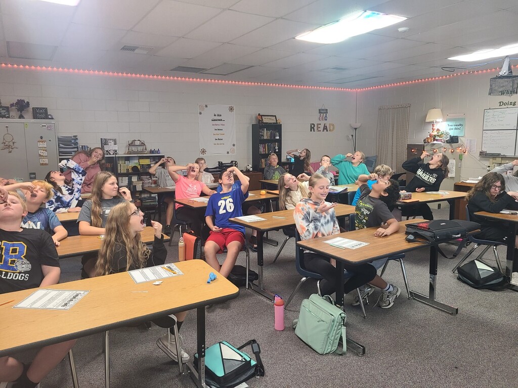 Sixth-grade students participate in a classroom activity, sitting at their desks and looking upward as they attempt to catch or balance small pieces of candy or food in their mouths.
