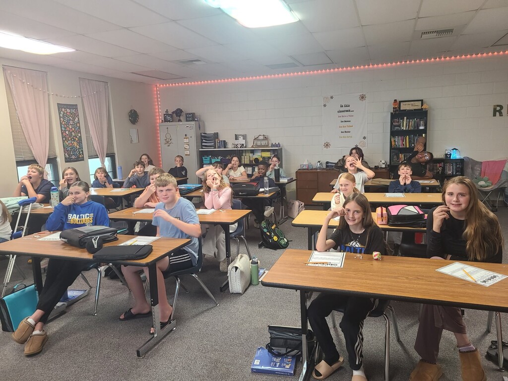Sixth-grade students sit at their desks during a classroom activity, smiling and holding small pieces of candy or food as they prepare to participate in a class challenge.