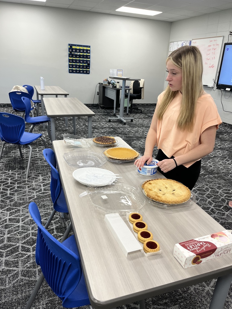 A student stands beside a classroom table preparing a selection of pies, tarts, plates, and other items for a class activity.